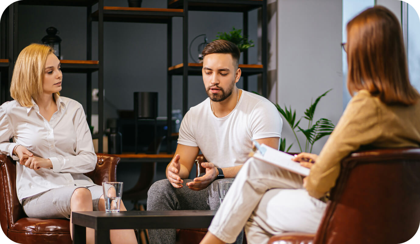 A couple sits in front of their therapist. The man speaks and gesticulates as the woman practices active listening, a couples therapy exercise that is helpful both in and out of session.