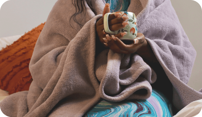 A female therapist sits with a blanket around her shoulders and a cup of hot tea as she thinks about overcoming barriers to self-care